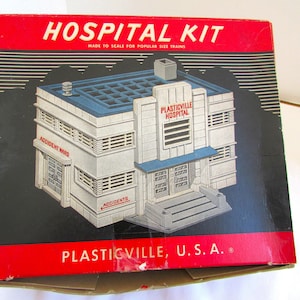 Könnte beinhalten: Eine Vintage-Krankenhaus-Kit-Box mit rotem und schwarzem Design. Die Box zeigt ein Modellkrankenhaus mit dem Text "PLASTICVILLE HOSPITAL" und "ACCIDENT WARD". Die Box ist mit "MADE TO SCALE FOR POPULAR SIZE TRAINS" und "PLASTICVILLE, U.S.A." beschriftet.