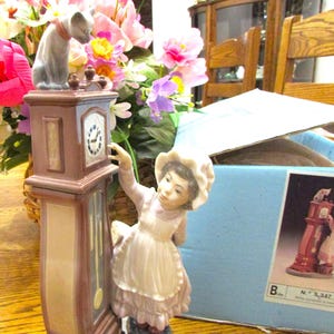 May include: A decorative figurine featuring a girl reaching for a grandfather clock, with a cat perched on top. The clock is brown and blue, and the girl wears a pink dress and white apron. A box with a picture of the figurine is in the background.