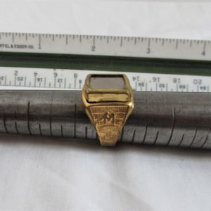 以下が含まれることがあります： 中央に長方形の薄い灰色の石がセットされた金の指輪。指輪は、前面に隆起した文字「M」が刻まれたテクスチャ加工されたバンドです。