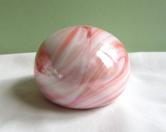 May include: A round, decorative glass paperweight with a swirling pattern of pink and white. The smooth, polished surface reflects light, creating highlights. The paperweight is set against a white cloth background.