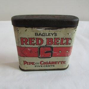 May include: Vintage tin container for Bagley's Red Belt Pipe or Cigarette tobacco. The tin is white with red and black accents and features a red belt with a buckle. The text "Bagley's Red Belt" is printed in black letters on the front of the tin. The text "Pipe or Cigarette Five Cents" is printed in black letters on the bottom of the tin.