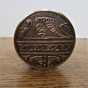 Puede incluir: Una perilla de puerta redonda de color cobre con un diseño intrincado en relieve. El diseño presenta un patrón floral estilizado y un motivo geométrico.