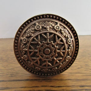 May include: A round, ornate, brass door knob with a detailed, circular design. The knob has a beaded edge and a raised, intricate pattern.
