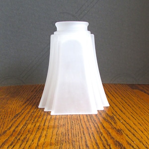 May include: A frosted white glass lamp shade with a square base and a flared, four-sided design. The shade features a smooth, matte finish and a circular opening at the top, tapering outwards from the neck to the wider, square-shaped bottom edge.
