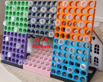 May include: A multi-colored storage rack featuring green, dark gray, orange, purple, light blue, and pink sections with circular cutouts. The rack holds numerous small, clear, cylindrical containers. The text "In 70 Farben" is printed in red.
