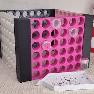 Könnte beinhalten: Ein rosa-schwarzer Aufbewahrungs-Organizer mit mehreren kreisförmigen Öffnungen für kleine Behälter. Der Organizer befindet sich neben einer weißen Schale mit kleinen, dunklen Perlen und einem rot-weißen Leuchtturm-förmigen Behälter.