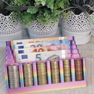 Könnte beinhalten: Ein mehrfarbiger Münz- und Geldschein-Organizer mit Farbverlauf, der sechs vertikale Fächer für gestapelte Münzen und drei horizontale Fächer für Geldscheine bietet. Der Organizer zeigt einen regenbogenartigen Übergang von violetten, blauen, gelben und rosa Farbtönen.