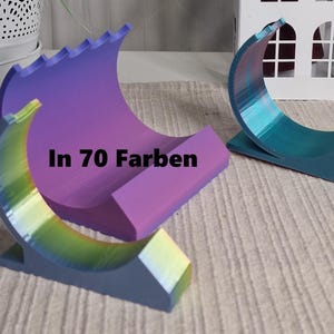 Op de afbeelding: Twee 3D-geprinte telefoonstandaards, een in een paars-blauwe gradiënt en de andere in een turquoise-blauwe gradiënt. De standaards zijn halfrond en hebben een getextureerd oppervlak. De tekst "In 70 Farben" is gedrukt op de paars-blauwe standaard.