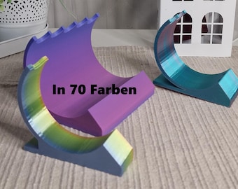 May include: Two 3D printed phone stands, one in a purple and blue gradient and the other in a teal and blue gradient. The stands are shaped like a crescent moon and have a textured surface. The text "In 70 Farben" is printed on the purple and blue stand.