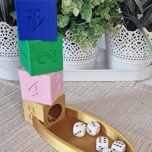 Op de afbeelding: Een gouden dobbelsteenbakje met een stapel gekleurde blokken. De blokken zijn blauw, groen en roze. Het blauwe blok heeft een symbool erop, het groene blok heeft het nummer 2 erop en het roze blok heeft het nummer 4 erop. Er zitten vijf witte dobbelstenen met zwarte stippen in het bakje.