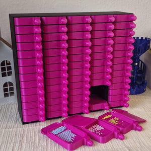 Könnte beinhalten: Ein magentafarbenes Aufbewahrungsmodul mit mehreren ausziehbaren Schubladen, ideal zum Organisieren kleiner Bastelutensilien. Das Modul hat einen schwarzen Rahmen und mehrere offene Schalen mit Perlen. Die Schalen sind geöffnet und zeigen den Inhalt.