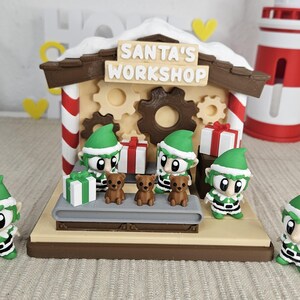 Könnte beinhalten: Ein festliches Diorama von Santa's Werkstatt mit Elfenfiguren, Teddybären und verpackten Geschenken. Die Werkstatt hat ein schneebedecktes Dach und den Text "SANTA'S WORKSHOP". Ein rot-weißer Leuchtturm befindet sich im Hintergrund.