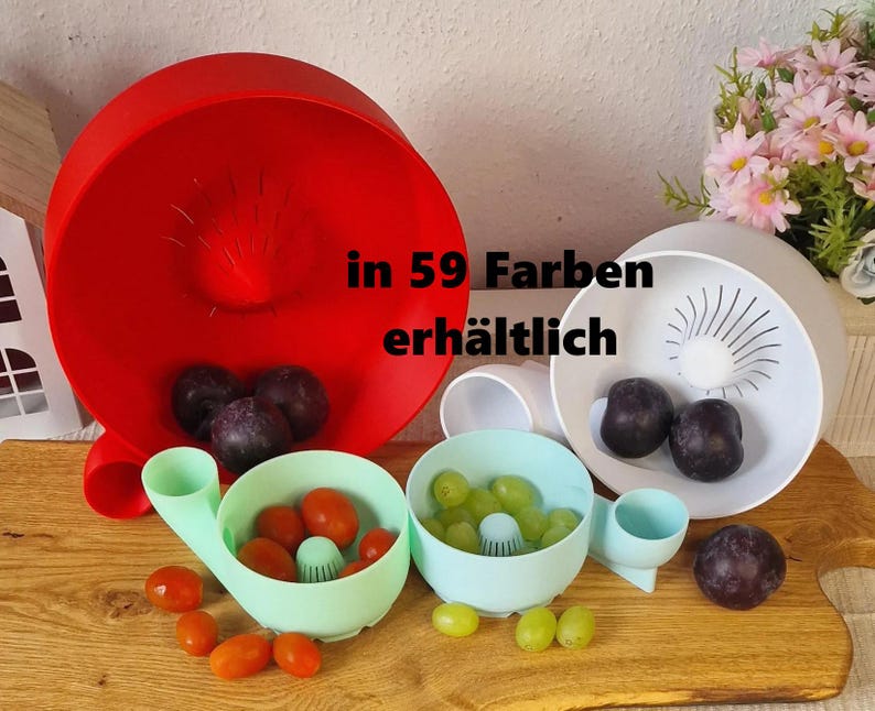 May include: A collection of colorful fruit washing bowls in red, white, and light blue, with a wooden cutting board base. The bowls contain grapes and tomatoes. The text "in 59 Farben erh&auml;ltlich" is visible.