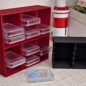 Könnte beinhalten: Ein roter Aufbewahrungs-Organizer mit mehreren Fächern und transparenten Kunststoffschubladen. Daneben befindet sich ein kleinerer schwarzer Organizer. Vor den Organisatoren befindet sich ein Behälter mit blauen Perlen. Im Hintergrund ein rot-weißes Objekt in Leuchtturmform.