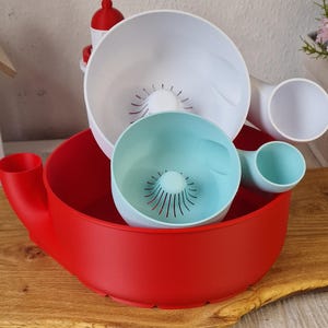 May include: A set of colorful kitchen tools, including a large red bowl with a handle, a white bowl, and a smaller teal bowl. The bowls have a unique design with drainage holes.