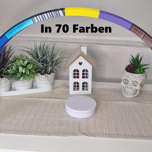Könnte beinhalten: Ein bunter Regenbogenbogen aus bemaltem Holz mit schwarzen Sockeln. Der Bogen ist mit Streifen in verschiedenen Farben verziert, darunter Blau, Gelb, Lila, Braun und Rosa. Der Bogen steht auf einem Tisch mit einer weißen runden Box davor. Der Text "In 70 Farben" ist auf dem Bogen geschrieben.