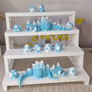 Può includere: Una mensola bianca a tre livelli espone statuette di draghi blu e uova bianche con pois blu. I draghi sono azzurri con dettagli bianchi. Le uova sono di varie dimensioni e disposizioni, creando un tema fantastico.