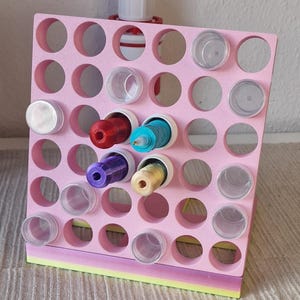 Op de afbeelding: Een roze schuimorganizer met meerdere cirkelvormige gaten, waarvan sommige spoelen met kleurrijk garen bevatten. Doorzichtige plastic containers zitten ook in sommige gaten. Bovenop staat een rood en wit object. De organizer heeft een veelkleurige basis.