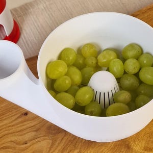 May include: A white plastic fruit washing and draining bowl filled with green grapes. The bowl has a built-in strainer and a spout. A red and white object is visible in the background. The bowl is on a wooden surface.