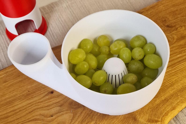 May include: A white plastic fruit washing and draining bowl filled with green grapes. The bowl has a built-in strainer and a spout. A red and white object is visible in the background. The bowl is on a wooden surface.