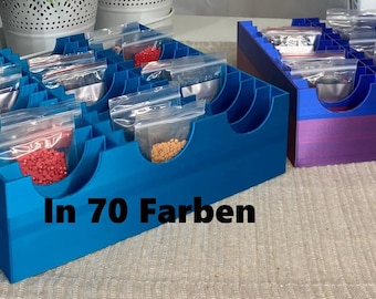 Puede incluir: Dos cajas de almacenamiento de plástico azul con múltiples compartimentos para artículos pequeños. Las cajas están llenas de pequeñas bolsas de cuentas de colores. El texto "In 70 Farben" está impreso en la parte delantera de la caja azul.
