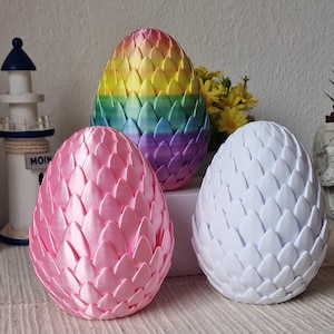 Könnte beinhalten: Drei dracheneierförmige Ornamente, eines ist rosa, eines ist weiß und eines ist regenbogenfarben. Das Regenbogen-Ei hat gelbe, orange, rote, lila und blaue Streifen.