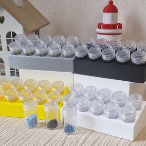 Op de afbeelding: Een verzameling kleine, transparante plastic potjes met schroefdeksels, gerangschikt op gekleurde, trapsgewijze displayblokken. De blokken zijn grijs, zwart, geel en wit. Sommige potjes bevatten kleine, gekleurde kralen.