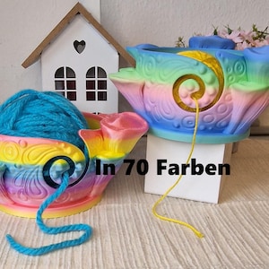 Può includere: Due ciotole per filato arcobaleno, ciascuna con filato. Le ciotole hanno un design a spirale e sono nelle tonalità di rosa, giallo, blu e verde. Il testo "In 70 Farben" è visibile.