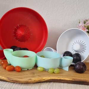 May include: A still life featuring a red colander, a white bowl, and two mint green bowls with attached spouts. The bowls are filled with fresh produce, including plums, grapes, and tomatoes. The items are arranged on a wooden cutting board.
