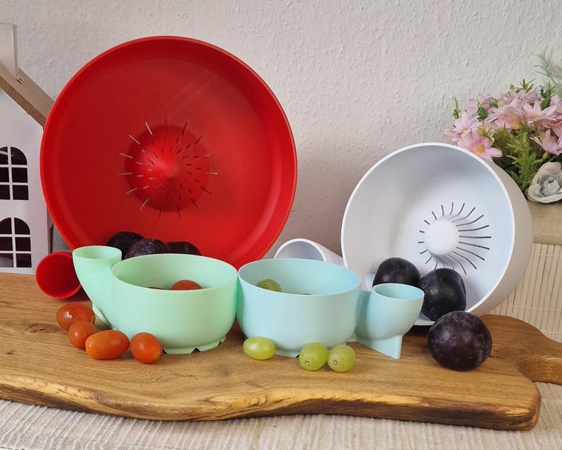 May include: A still life featuring a red colander, a white bowl, and two mint green bowls with attached spouts. The bowls are filled with fresh produce, including plums, grapes, and tomatoes. The items are arranged on a wooden cutting board.