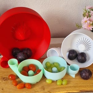 May include: A collection of colorful kitchen colanders and bowls. A large red colander is in the background, with a white and two light blue bowls in the foreground. The bowls contain grapes and tomatoes. The colanders are on a wooden cutting board.