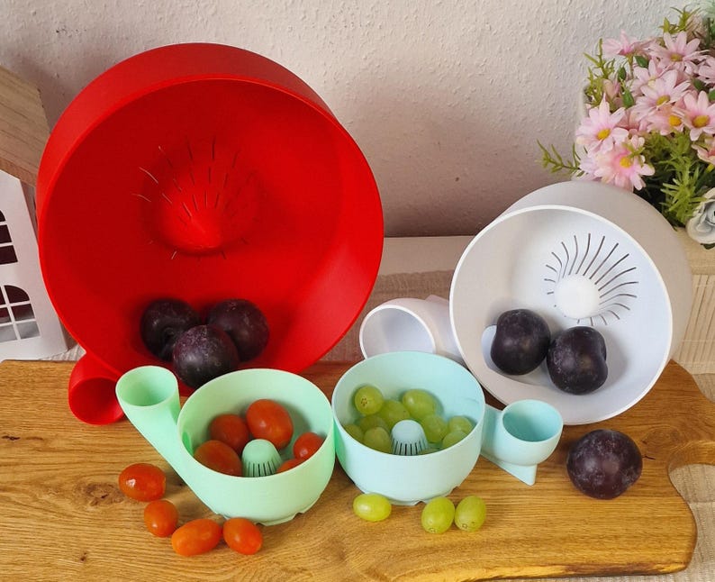 May include: A collection of colorful kitchen colanders and bowls. A large red colander is in the background, with a white and two light blue bowls in the foreground. The bowls contain grapes and tomatoes. The colanders are on a wooden cutting board.