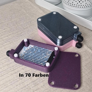 Könnte beinhalten: Eine violette und rosafarbene Plastik-Aufbewahrungsbox mit einem durchsichtigen Deckel. Der Deckel hat ein Gitter aus kleinen blauen Quadraten. Die Box hat den Text "In 70 Farben" auf der Seite.