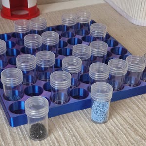 Könnte beinhalten: Ein blauer Kunststoffbehälter mit zahlreichen zylindrischen, transparenten Kunststofffläschchen mit Schraubdeckeln. Einige Fläschchen enthalten kleine, dunkle Perlen, andere blaue Granulate. Im Hintergrund ist eine rot-weiße Leuchtturmdekoration zu sehen.