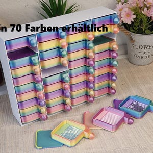 Könnte beinhalten: Ein weißer Aufbewahrungsorganizer mit mehreren Fächern, der zahlreiche kleine, quadratische, regenbogenfarbene Schalen mit kugelförmigen Knöpfen enthält. Der Text "in 70 Farben erhältlich" ist oben links aufgedruckt.
