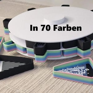 Puede incluir: Un carrusel de almacenamiento giratorio blanco con 12 compartimentos para artículos pequeños. Los compartimentos están forrados con silicona arcoíris. El carrusel está etiquetado "In 70 Farben".