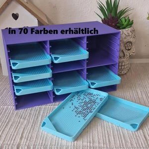 Könnte beinhalten: Ein violetter Aufbewahrungsorganizer mit mehreren Fächern für hellblaue Schalen. Die Einheit verfügt über horizontale Regale zur Aufnahme der Schalen, wobei zwei Schalen davor platziert sind. Der Text "in 70 Farben erhältlich" ist oben auf der Einheit aufgedruckt.