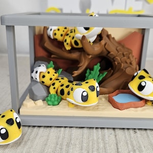 Op de afbeelding: Een grillig diorama met geel en zwart gevlekte gekko-beeldjes. De scène omvat een grijze kooi, een bruine boomstam, groene planten en een kleine waterbak. Eén gekko bevindt zich buiten de kooi.