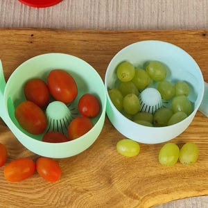 May include: Two light blue kitchen tools, each with a bowl and handle, are filled with red tomatoes and green grapes. The tools sit on a wooden surface, with loose tomatoes and grapes scattered around.
