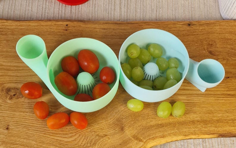 May include: Two light blue kitchen tools, each with a bowl and handle, are filled with red tomatoes and green grapes. The tools sit on a wooden surface, with loose tomatoes and grapes scattered around.