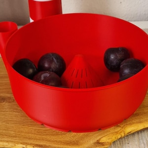 May include: A bright red fruit washing bowl with a built-in strainer and a handle. The bowl contains several dark purple plums. The bowl is sitting on a wooden cutting board.