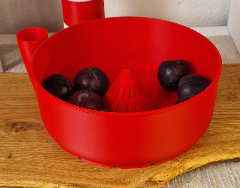 May include: A bright red fruit washing bowl with a built-in strainer and a handle. The bowl contains several dark purple plums. The bowl is sitting on a wooden cutting board.