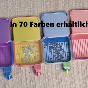 May include: Four open, colorful plastic containers with lids, each holding small, shiny objects. The containers are purple, yellow, blue, and pink. The text "in 70 Farben erhältlich" is visible.