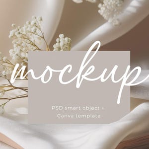 May include: A mockup card with the word "mockup" written in white cursive script on a light gray background. The text "PSD smart object + Canva template" is written in black below the word "mockup". The card is lying on a white fabric with white flowers.