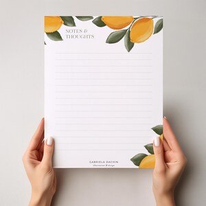 May include: A white notepad with lined pages, held by hands. The notepad features watercolor illustrations of lemons and leaves, with the text "NOTES & THOUGHTS" at the top and the designer's name at the bottom.