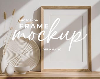 May include: A wooden framed mockup with the text "Photoshop Frame Mockup" in white script. The frame is empty, with a white mount. Decorative white ceramic plate, vase, and bowl are on a wooden surface.