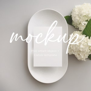 Puede incluir: Maqueta de tarjeta blanca en un plato ovalado blanco con hortensias blancas sobre un fondo gris. La tarjeta tiene el texto "mockup" escrito en cursiva y el texto "PSD smart object + Canva template".