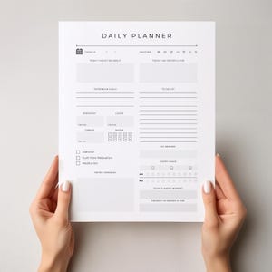 May include: A white daily planner with black text, held by hands. The planner includes sections for goals, to-do lists, and tracking habits. The text "DAILY PLANNER" is at the top. Sections include "Today I'm Excited About" and "Today I Am Grateful For."