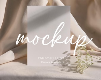 Puede incluir: Una maqueta de tarjeta blanca con la palabra "mockup" escrita en cursiva. La tarjeta está sobre una superficie de tela marrón claro con un pequeño ramo de flores blancas en primer plano. El texto "PSD smart object + Canva template" está impreso en la tarjeta.