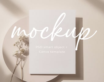 May include: A white card mockup with the word "mockup" written in cursive on a beige background. The card is sitting on a beige plate with a sprig of white flowers.